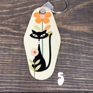May include: A cream-colored keychain with a retro design featuring a black cat and orange flowers. The cat has large eyes and a long tail. The number "5" is below the keychain. A silver key ring is attached.