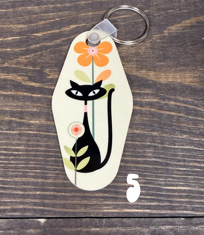 May include: A cream-colored keychain with a retro design featuring a black cat and orange flowers. The cat has large eyes and a long tail. The number "5" is below the keychain. A silver key ring is attached.