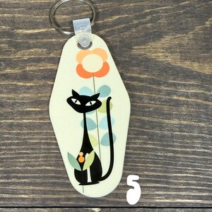 May include: A retro-style keychain featuring a black cat with a long tail, a flower, and a light green background. The keychain has a metal ring and a plastic clasp. The number "5" is visible on the bottom right.