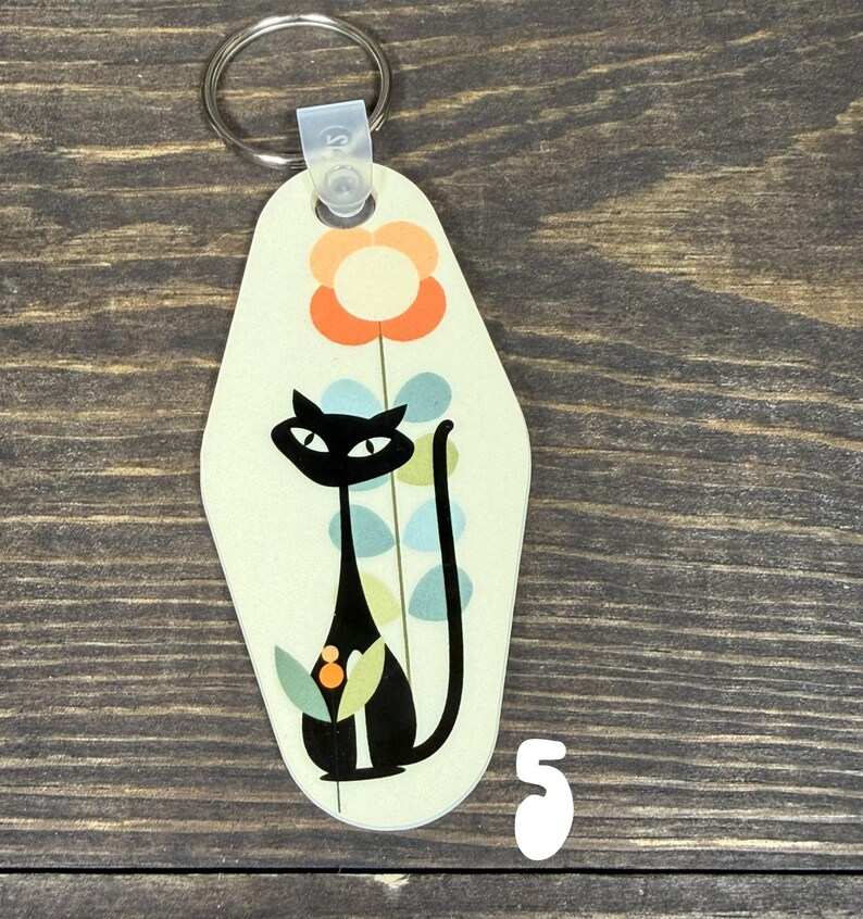 May include: A retro-style keychain featuring a black cat with a long tail, a flower, and a light green background. The keychain has a metal ring and a plastic clasp. The number "5" is visible on the bottom right.