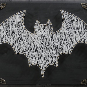 May include: White string art bat design on a black background. The bat is created with white string wrapped around nails hammered into the wood. The bat is centered on the black background.
