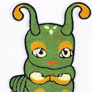 May include: A green plush caterpillar with yellow and orange accents. The caterpillar has a smiling face with large eyes and a small mouth. It has its arms crossed in front of it.