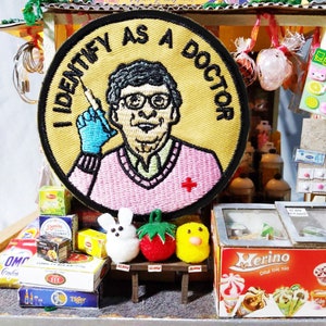 May include: A miniature shop with a yellow and black embroidered patch that says "I Identify as a Doctor". The patch features a cartoon image of a person wearing a pink sweater and holding a syringe. The shop also has a variety of other items, including a small box of ice cream, a box of cookies, and a few other small items.