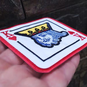 May include: Red and white embroidered patch with a king of hearts playing card design. The king is wearing a gold crown and has a blue face.