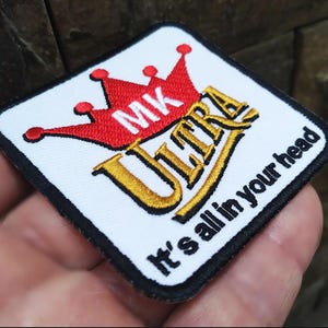Può includere: Toppa ricamata bianca con una corona rossa e lettere dorate che dicono "MK Ultra" e "It's all in your head".