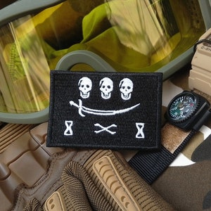 Op de afbeelding: Zwart-witte geborduurde patch met drie schedels, een kromzwaard, gekruiste botten en zandlopers. De patch is op een camouflage achtergrond met een bril, een horloge en handschoenen.