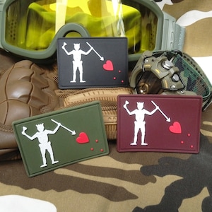 Könnte beinhalten: Drei gestickte Patches mit einem weißen Skelett, das eine Lanze und ein rotes Herz hält. Die Patches befinden sich auf einem Tarnmuster-Hintergrund. Die Patches sind schwarz, grün und rotbraun.