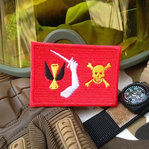 Pode incluir: Um remendo bordado vermelho com um braço branco segurando uma espada, um pássaro preto com um bico amarelo e uma caveira amarela e ossos cruzados. O remendo está sobre um fundo de couro marrom.