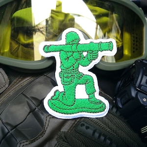 Könnte beinhalten: Grüner gestickter Aufnäher mit einem Spielzeugsoldaten, der einen Raketenwerfer hält. Der Aufnäher ist auf weißem Hintergrund und hat einen weißen Rand.