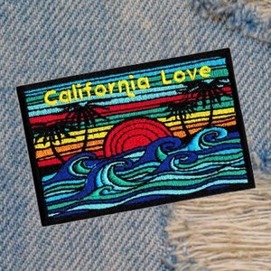 Może przedstawiać: Prostokątna naszywka z żółtym napisem "California Love", przedstawiająca scenę zachodu słońca z palmami, czerwonym słońcem i niebieskimi falami. Naszywka znajduje się na tle zniszczonego dżinsu.