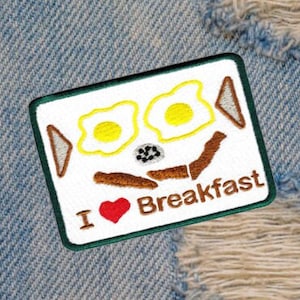 Puede incluir: Un parche bordado blanco con un borde verde. El parche presenta una imagen de estilo de dibujos animados de dos huevos amarillos, salchichas marrones y el texto "Me encanta el desayuno".