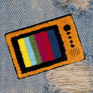 May include: A yellow embroidered patch of a vintage television with a colorful static screen. The patch is on a blue denim background.
