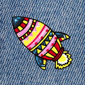 Può includere: Una toppa a forma di razzo ricamata colorata con punta nera, dettagli rosa, gialli, rossi e blu. Il razzo ha un design dettagliato ed è posto su uno sfondo di denim blu.