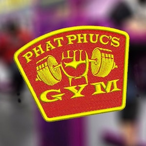 Puede incluir: Un parche rojo y amarillo con el texto "PHAT PHUC'S GYM" en letras amarillas. El parche presenta un gráfico amarillo de una mano levantando una barra con pesas en cada extremo.