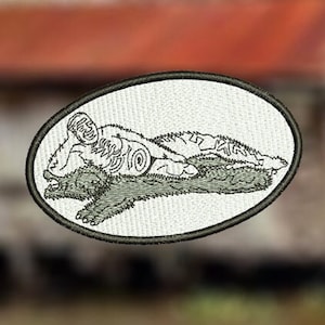 Może przedstawiać: Biała haftowana łatka z czarno-białym obrazkiem osoby leżącej na dużym zwierzęciu. Osoba ma na sobie kapelusz, a zwierzę ma długi ogon.