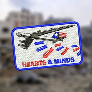 以下が含まれることがあります： 青い縁取りのある白いパッチには、赤と青のストライプの爆弾を落とす爆撃機の漫画が描かれています。飛行機の下には「HEARTS & MINDS」という文字が印刷されています。