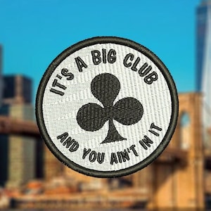 Może przedstawiać: Naszywka haftowana w kolorze białym i czarnym z tekstem "It's a big club and you ain't in it" i czarnym symbolem koniczyny. Naszywka znajduje się przed rozmytym tłem miejskiego krajobrazu i mostu.