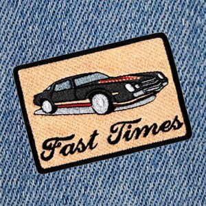 Puede incluir: Un parche bordado beige con un coche negro y el texto "Fast Times" en cursiva negra.