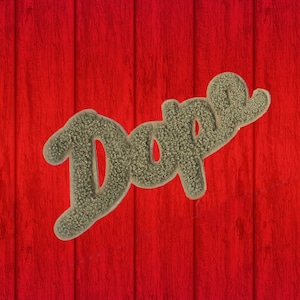 Puede incluir: La palabra "Dope" en una fuente texturizada de color marrón claro está centrada en un fondo de madera roja. La veta de la madera corre verticalmente y el color rojo tiene un efecto degradado, siendo el centro más claro.