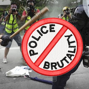 May include: A protester holds a sign that reads "No Police Brutality" in red lettering with a diagonal red line through the words. The sign is in front of a police officer holding a riot shield. The officer is wearing a black helmet and a black uniform. The officer is standing in a street with other protesters and police officers in the background.