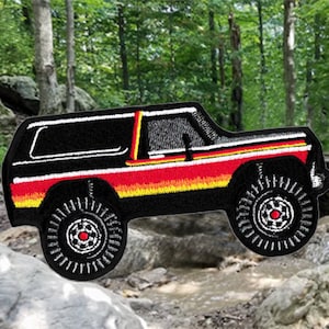 Op de afbeelding: Zwarte en rode geborduurde patch van een vintage terreinwagen met een zwarte achtergrond en gele, oranje en rode strepen. Het voertuig bevindt zich op een witte achtergrond.