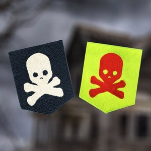 May include: Two fabric patches with skull and crossbones designs. The patch on the left is dark blue with a white skull and crossbones. The patch on the right is bright yellow with a red skull and crossbones.