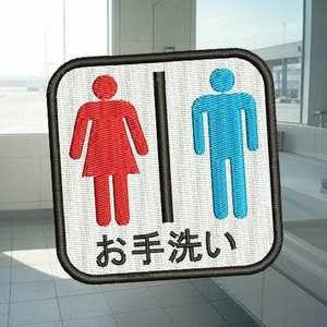 Puede incluir: Un letrero blanco con borde negro y fondo negro con el texto "お 手 洗 い" en blanco. El letrero tiene un símbolo femenino rojo y un símbolo masculino azul sobre fondo blanco. El letrero está en un baño con un lavabo blanco y paredes de azulejos blancos.