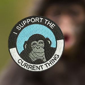 Puede incluir: Un parche bordado en blanco y negro con un círculo azul en el centro. El círculo presenta una imagen en blanco y negro de un chimpancé con una expresión seria. El texto alrededor del círculo dice "I Support The Current Thing".