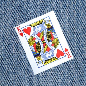 Peut inclure: Une carte à jouer avec une couleur de cœur rouge. La carte est le Roi de Cœur et présente un portrait d'un roi avec une couronne et une épée. La carte est sur un fond en jean bleu.