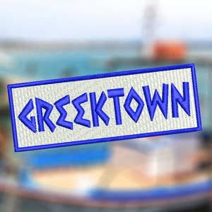 Peut inclure: Écusson brodé avec le mot "GREEKTOWN" en bleu, lettrage stylisé. Le patch a un fond blanc et une bordure bleue. L'arrière-plan est une image floue d'un bateau sur l'eau.