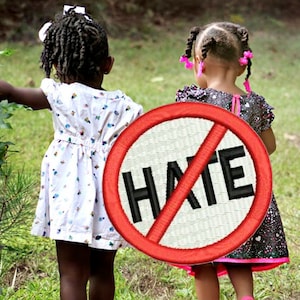 Puede incluir: Dos niñas pequeñas, una con un vestido blanco con lunares negros y la otra con un vestido negro y rosa, caminan lado a lado sobre un puente de madera. La niña con el vestido negro y rosa lleva un círculo rojo con una línea diagonal que lo atraviesa y la palabra "HATE" escrita en letras negras.