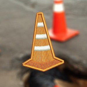 May include: A close-up of a small orange and white traffic cone patch. The patch is embroidered and has a stitched border. The patch is sitting on a gray asphalt surface with a large hole in the center.