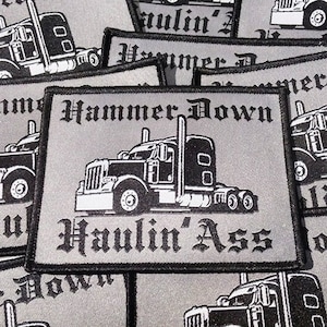 Puede incluir: Primer plano de varios parches bordados grises y negros con un diseño de camión de plataforma. Los parches muestran el texto "Hammer Down" y "Haulin' Ass".