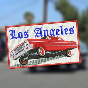 Pode incluir: Patch bordado com um carro lowrider vermelho e o texto "Los Angeles" em escrita azul. O patch tem um fundo branco e uma borda vermelha, representando um tema de cultura automotiva clássica.