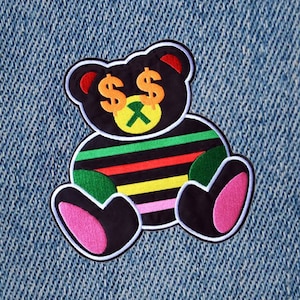 May include: A black and white teddy bear with dollar signs for eyes and a green, yellow, red, and pink striped shirt. The bear is sitting on a blue denim background.