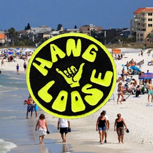 Puede incluir: Escena de playa soleada con un parche circular que muestra "HANG LOOSE" y un gesto con la mano. El parche es negro con letras amarillas. La gente disfruta de la playa, con edificios al fondo.