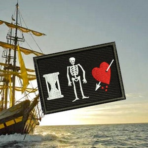 May include: A black pirate flag with a white hourglass, a skeleton, a red heart with an arrow through it, and three red drops of blood. The flag is on a ship sailing on the ocean.
