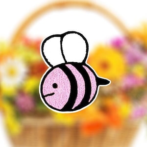 May include: A pink and black cartoon bee with white wings. The bee is in focus and the background is blurred, showing a basket of flowers.