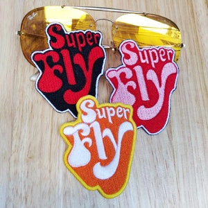 Puede incluir: Tres parches bordados para planchar con el texto "Super Fly" en una fuente retro. Los parches son de color rojo, rosa y naranja con letras blancas.
