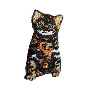 Op de afbeelding: Een zwart, oranje en wit kussen met pailletten in de vorm van een kat. De kat zit met zijn poten ingetrokken. Hij heeft blauwe ogen en een glimlachend gezicht.