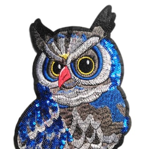 Puede incluir: Una aplicación de búho de lentejuelas azules y plateadas con patas rojas. El búho tiene ojos grandes y redondos con pupilas negras e iris amarillos.