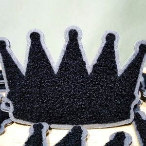Könnte beinhalten: Schwarzer Filz-Kronen-Patch mit grauem Umriss. Der Patch ist eine beliebte Verzierung für Kleidung, Taschen und andere Accessoires.