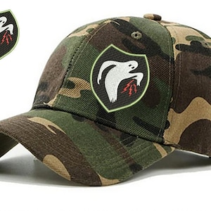 Puede incluir: Gorra de béisbol de camuflaje con un fantasma blanco que sostiene rayos bordados en la parte delantera. El fantasma está dentro de un escudo negro.