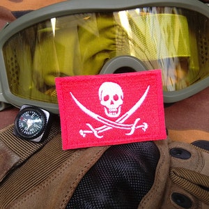 May include: A red fabric patch with a white skull and crossbones design. The patch is on a camouflage background with a pair of goggles and a compass.