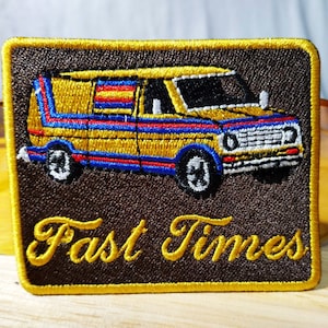 Puede incluir: Un parche bordado marrón y amarillo con una furgoneta amarilla y el texto "Fast Times".