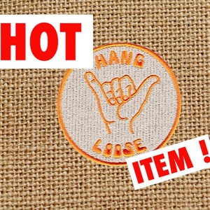 Puede incluir: Parche bordado naranja con una mano blanca haciendo el gesto de "hang loose". El parche está sobre un fondo de arpillera marrón. El texto "HANG LOOSE" está bordado en naranja alrededor de la mano. El texto "HOT ITEM!" está en rojo sobre fondo blanco.