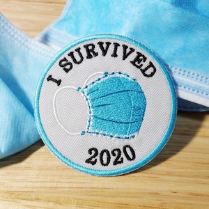 Op de afbeelding: Witte geborduurde patch met de tekst "I Survived 2020" en een blauwe gezichtsmasker afbeelding. De patch ligt op een houten oppervlak met twee blauwe gezichtsmaskers.