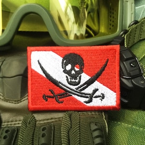 Puede incluir: Un parche de bandera de buceo rojo y blanco con un diseño de calavera y huesos cruzados negros. El parche está bordado y tiene un borde blanco.