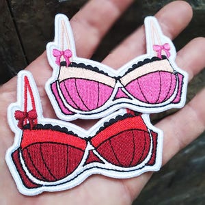 May include: Two embroidered iron-on patches in the shape of bras. One is pink and white, the other is red and white.