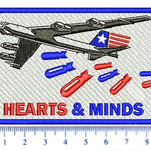 以下が含まれることがあります： 青い縁取りのある白い刺繍パッチ。パッチには、爆撃機が爆弾を投下している白黒のイラストが描かれています。爆弾は赤、白、青です。飛行機の下には「HEARTS & MINDS」という文字が赤と青の文字で書かれています。